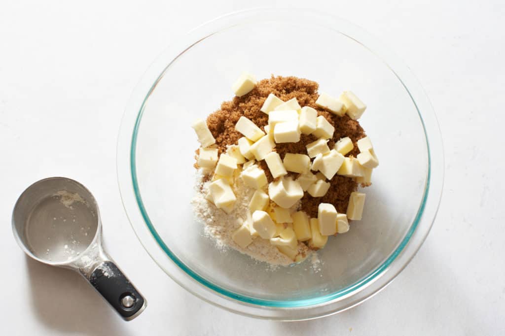A measuring cup next to a glass bowl filled with sugar, flour, and cubes of butter.