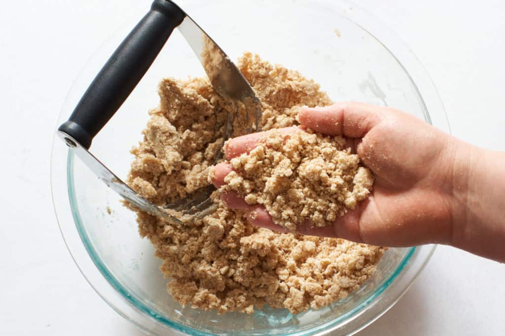 A woman's hand holding crumble topping over a bowl full of the same.