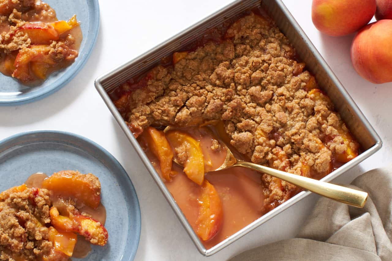 A square baking pan of peach crumble with a gold spoon in it surrounded by two blue plates of peach crumble, fresh peaches and a brown napkin.