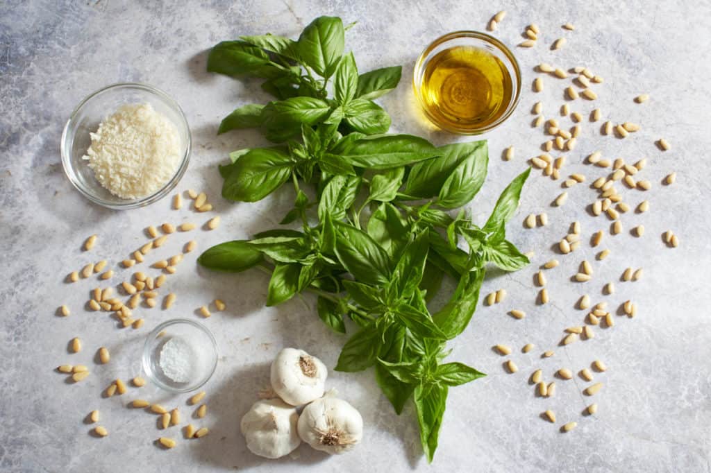 Pine nuts, fresh basil and garlic cloves with three small bowls of salt, parmesan cheese and olive oil.