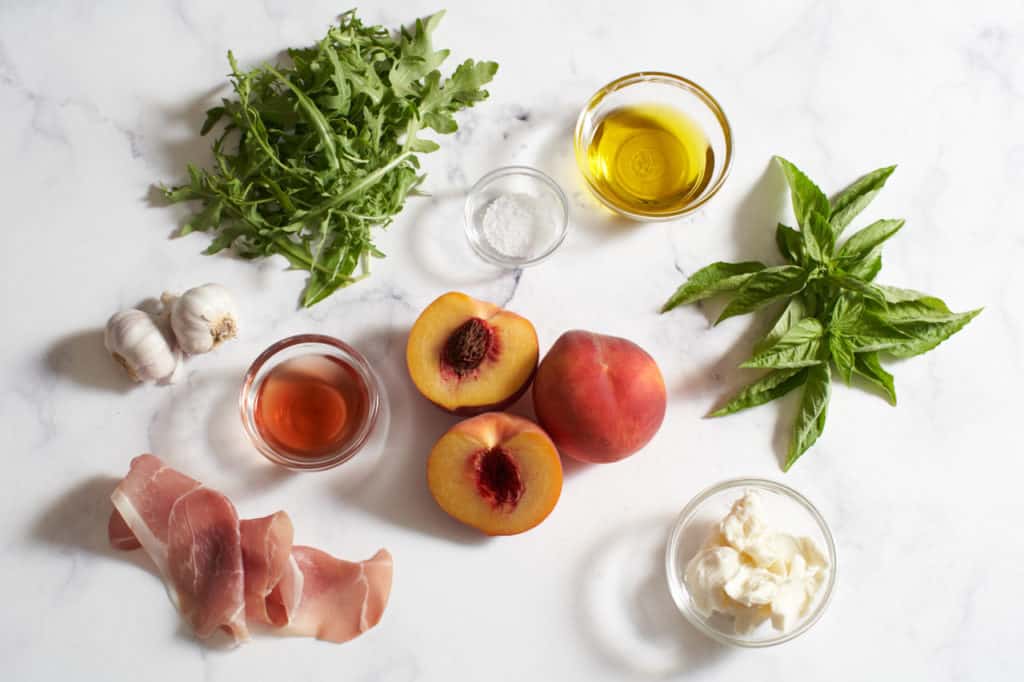 Arugula, basil, peaches, olive oil, red wine vinegar, garlic, prosciutto and fresh mozzarella on a marble surface.