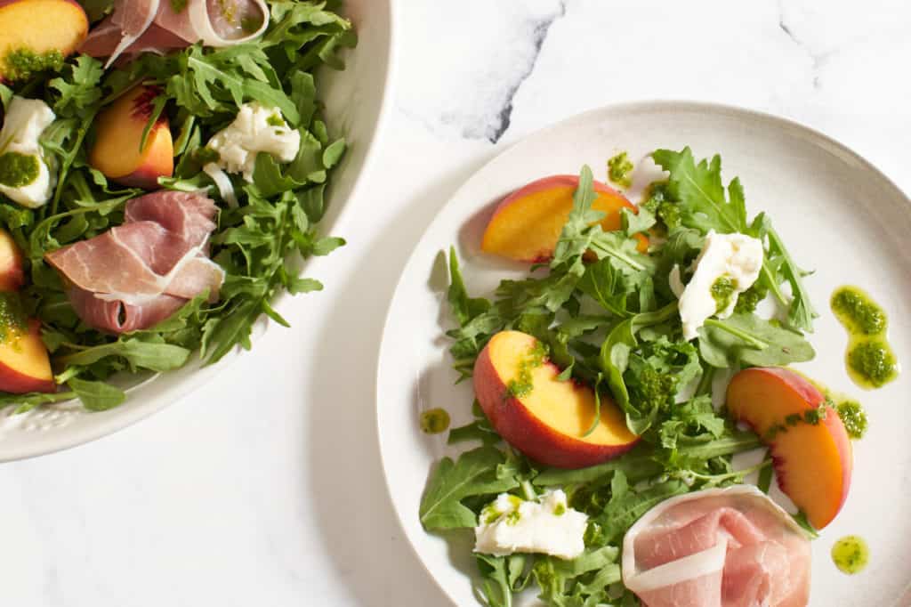 A large white bowl and a small white plate with peach and prosciutto salad.