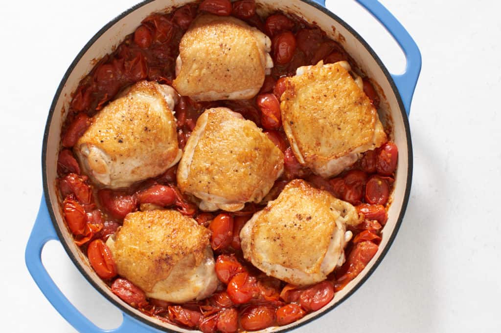 Browned chicken thighs atop burst cherry tomato sauce in a blue pan.