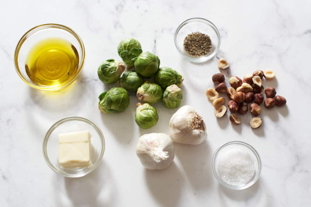 Brussels sprouts, hazelnuts, garlic bulbs, and small bowls with butter, olive oil, and salt and pepper.