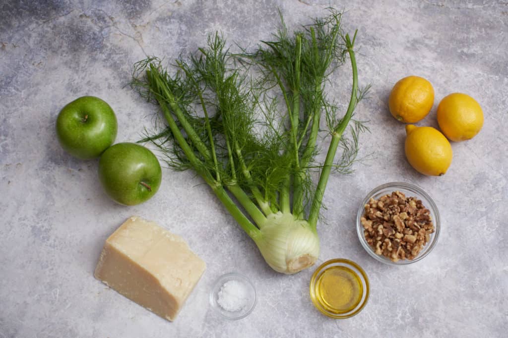 Apples, lemons, a fennel bulb, parmesan cheese, and small bowls with olive oil, salt, and walnuts.