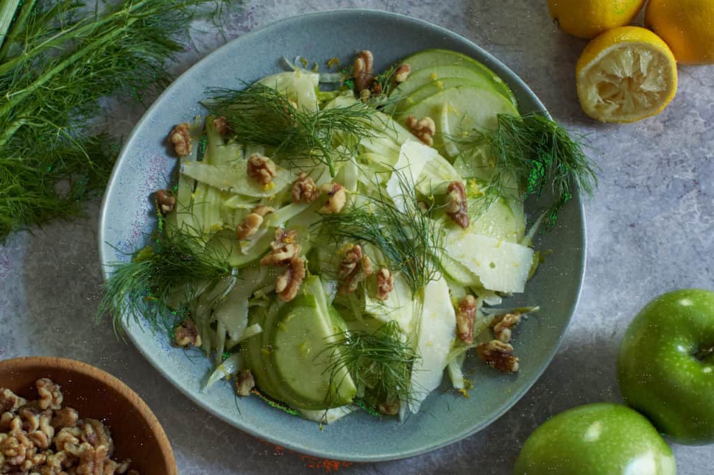 Apple fennel salad on a gray plate surrounded by lemons, apples, fennel fronds and walnuts.