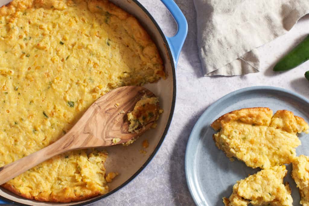 A casserole pan and a plate of spoonbread with a napkin and jalapeños in the background.