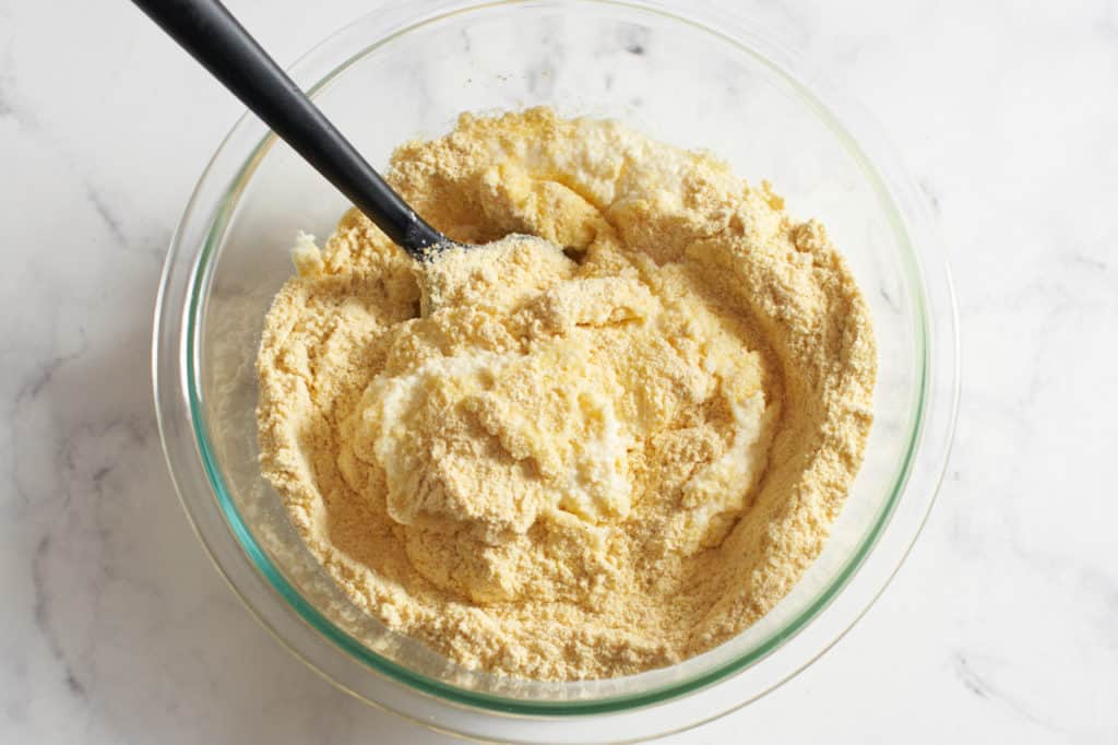 Cornmeal is added to a bowl of ricotta batter with a spatula in it.