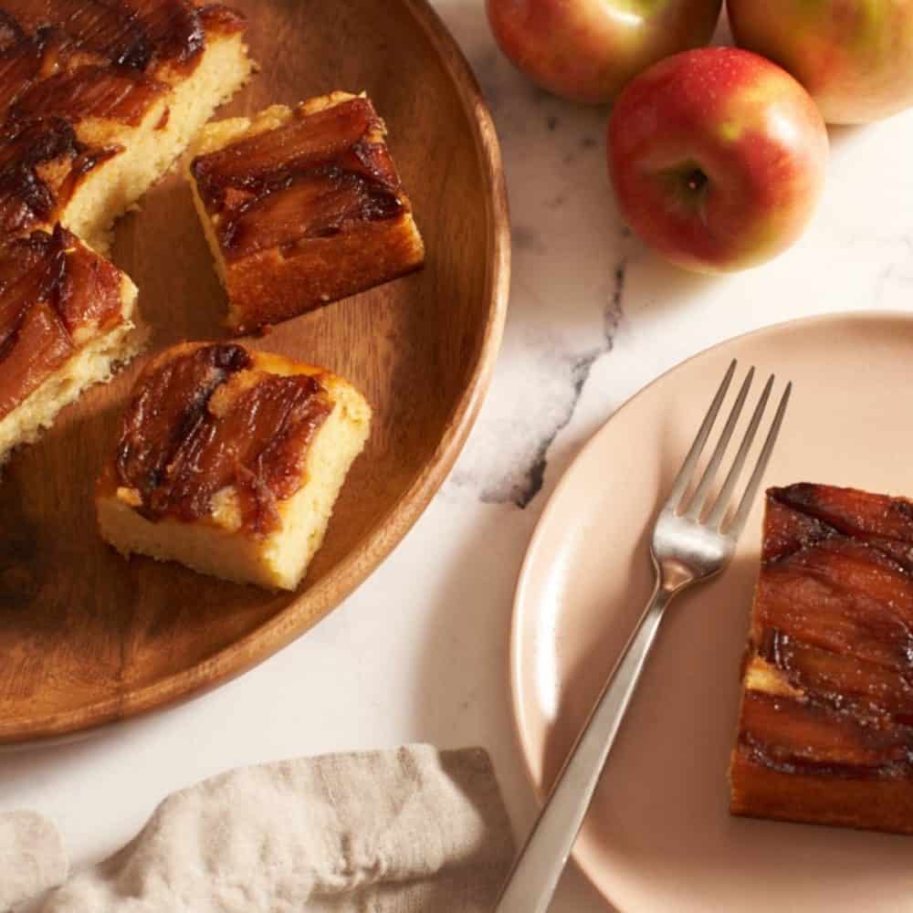 Apple ricotta cake on a wooden plate next to a small plate with a slice of cake on it.