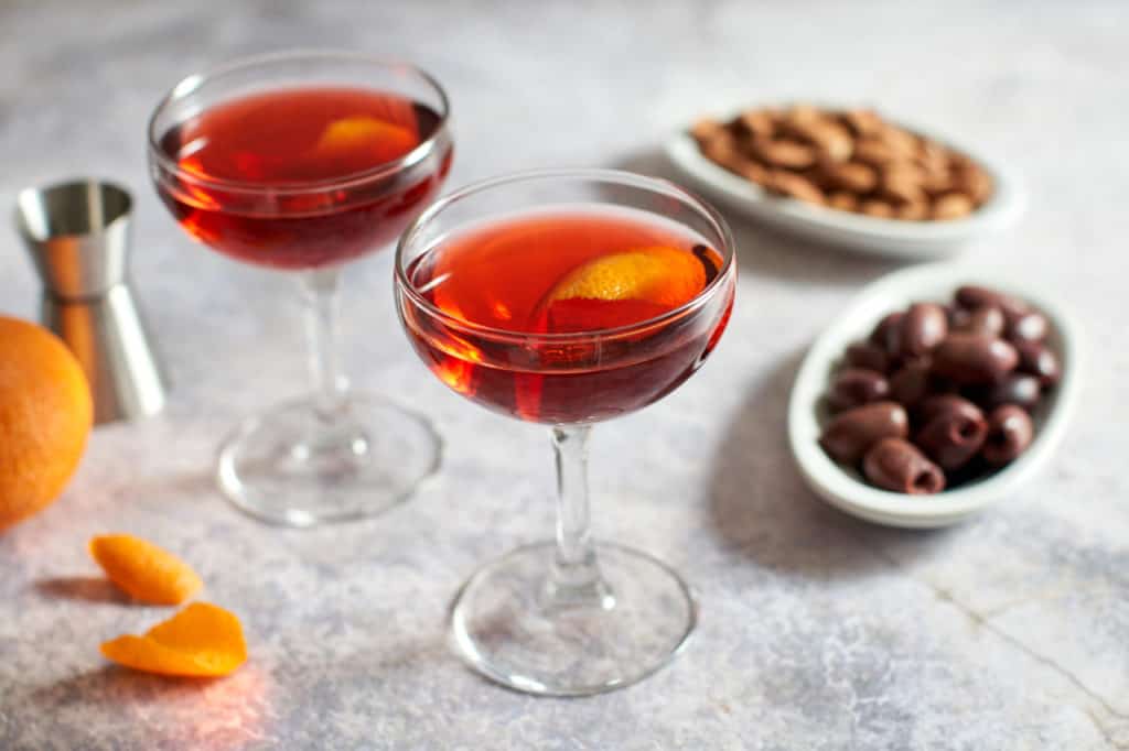 Two coupe glasses of Boulevardier cocktails, alongside small plates of almonds and olives. A cocktail jigger is in the background.