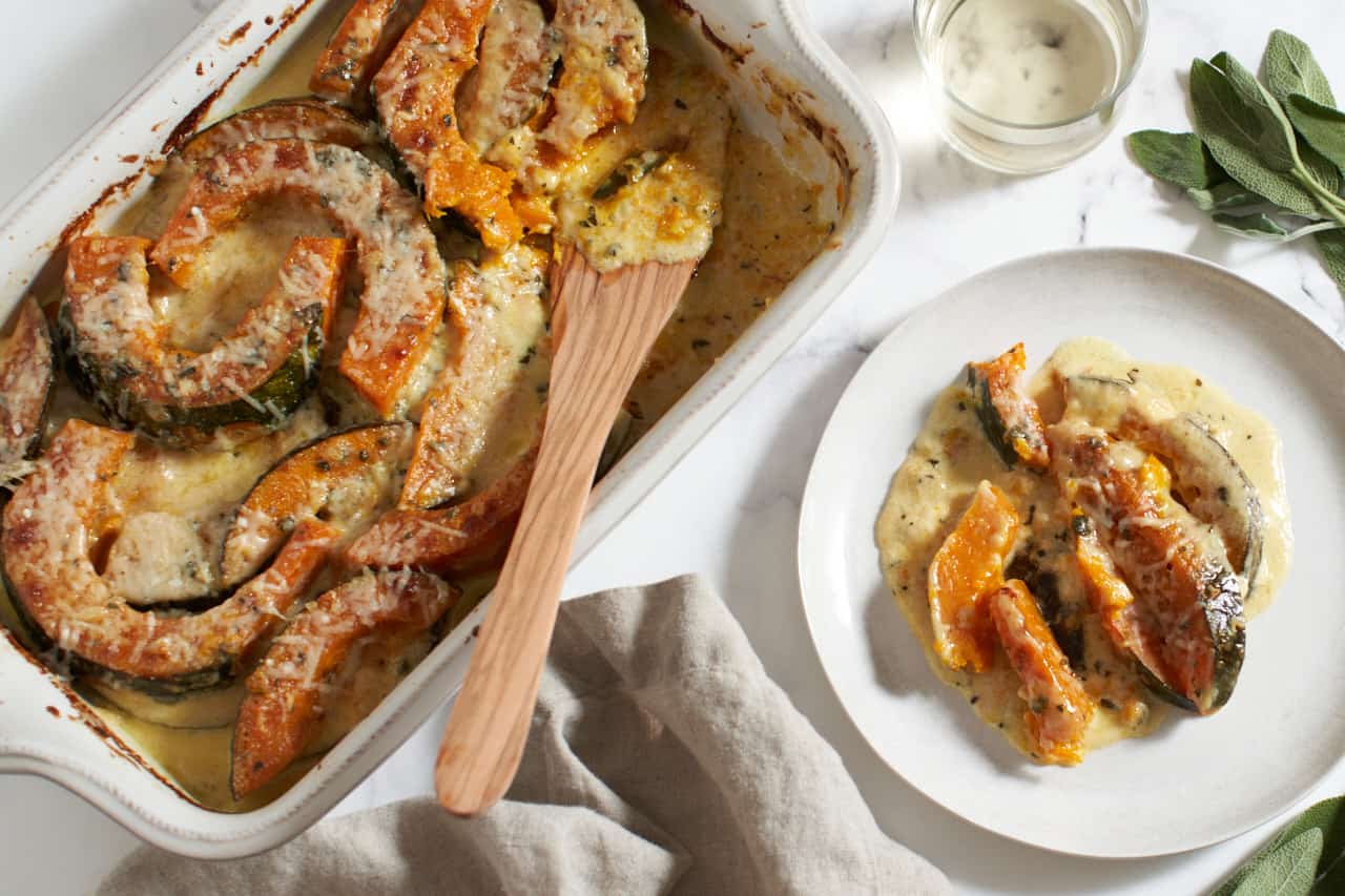 Winter squash gratin in a baking dish with a wooden spatula next to a plate of the same. A glass of white wine, a brown napkin, and sage leaves are nearby.