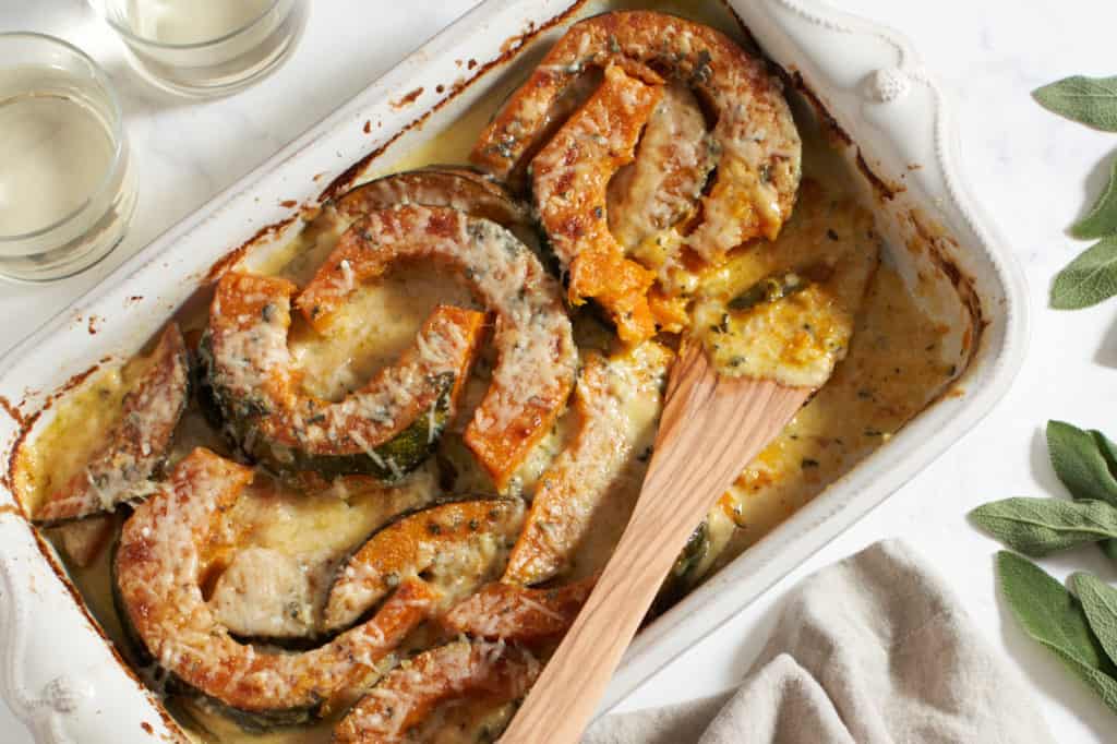 A baking dish of winter squash gratin with a wooden spatula surrounded by fresh sage leaves, a brown napkin, and a glass of wine.