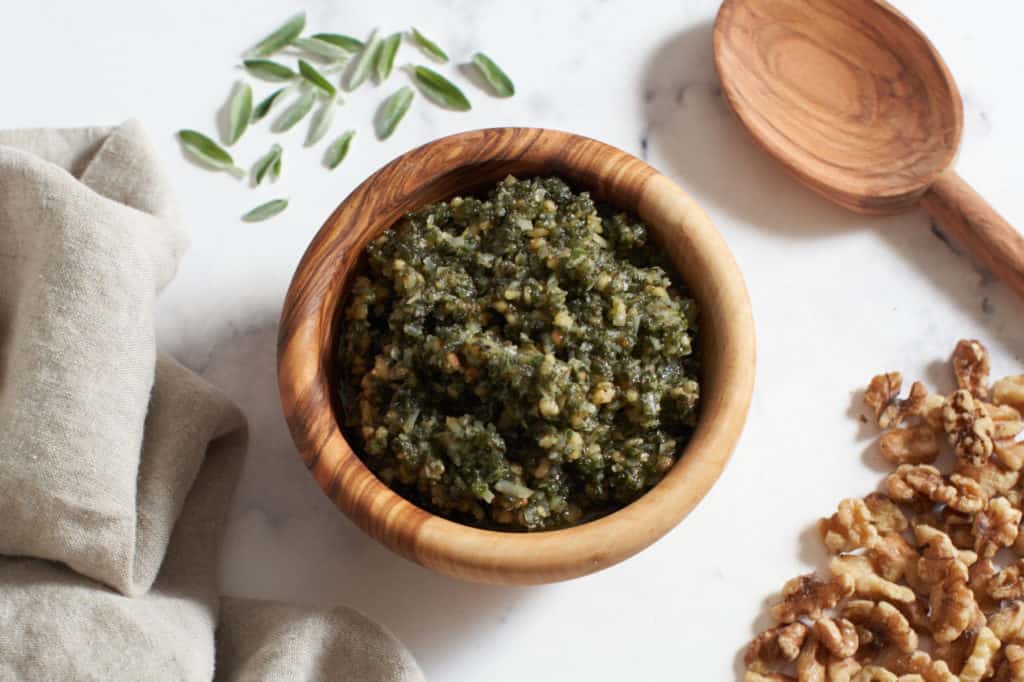 Sage pesto in a wooden bowl surrounded by walnuts, fresh sage leaves, a napkin, and a wooden spoon.