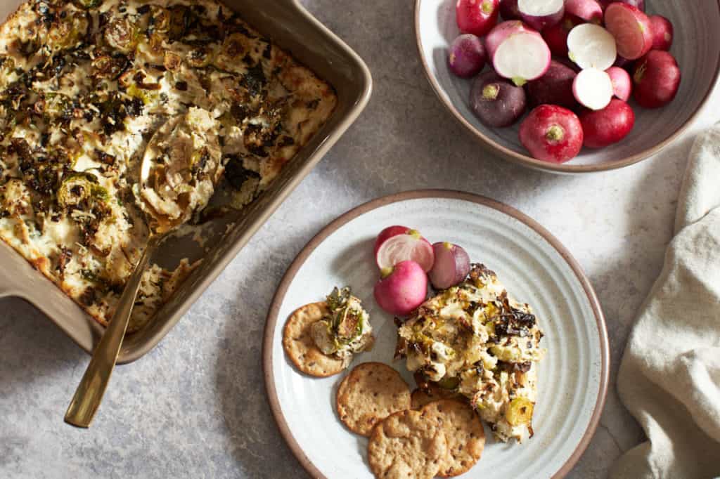 A brown pan of brussels sprouts dip next to a bowl of radishes and a small plate of dip and crackers.