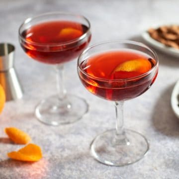 Two coupe glasses of Boulevardier cocktails, alongside small plates of almonds and olives.
