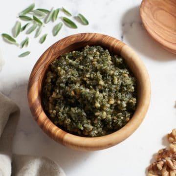 Sage pesto in a wooden bowl surrounded by walnuts, fresh sage leaves, a napkin, and a wooden spoon.