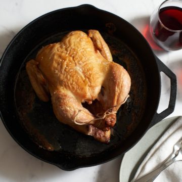 A roast chicken in a cast iron skillet with a glass of wine, two plates, forks and a napkin on the right.