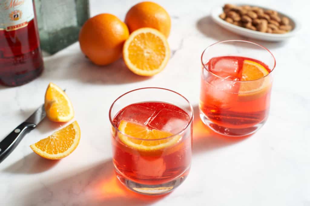 Two campari and soda cocktails with orange slices, a plate of nuts and two bottles in the background.