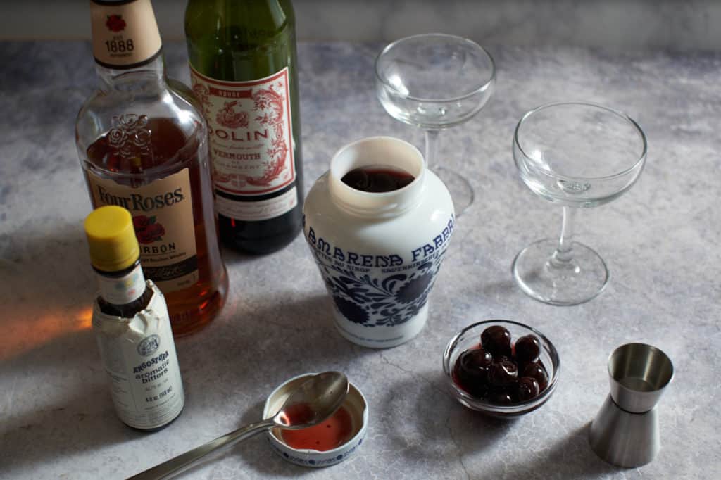 Bottles of bourbon, vermouth and bitters, with two coupe glasses, a jar and a bowl of cherries, and a cocktail jigger.