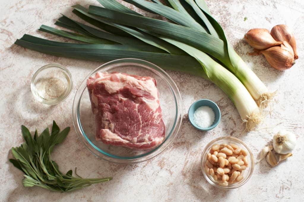 Pork shoulder in a glass bowl surrounded by shallots, garlic, sage, and small bowls with white beans, white wine and salt.