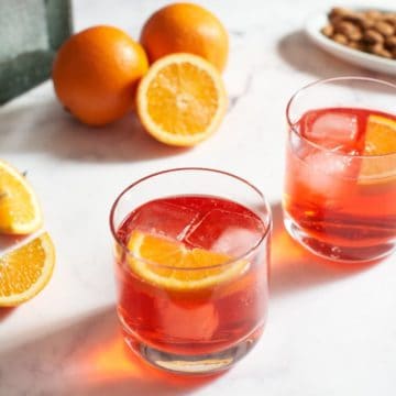 Two campari and soda cocktails with orange slices, a plate of nuts and two bottles in the background
