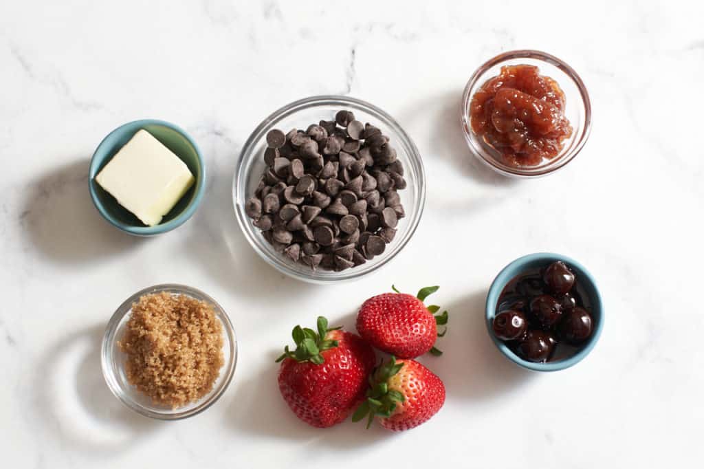 Three strawberries and small bowls of butter, brown sugar, chocolate chips, cherries, and jam.