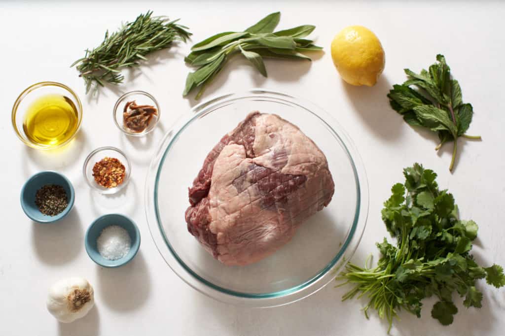 A raw boneless lamb leg surrounded by fresh herbs and small bowls of spices.