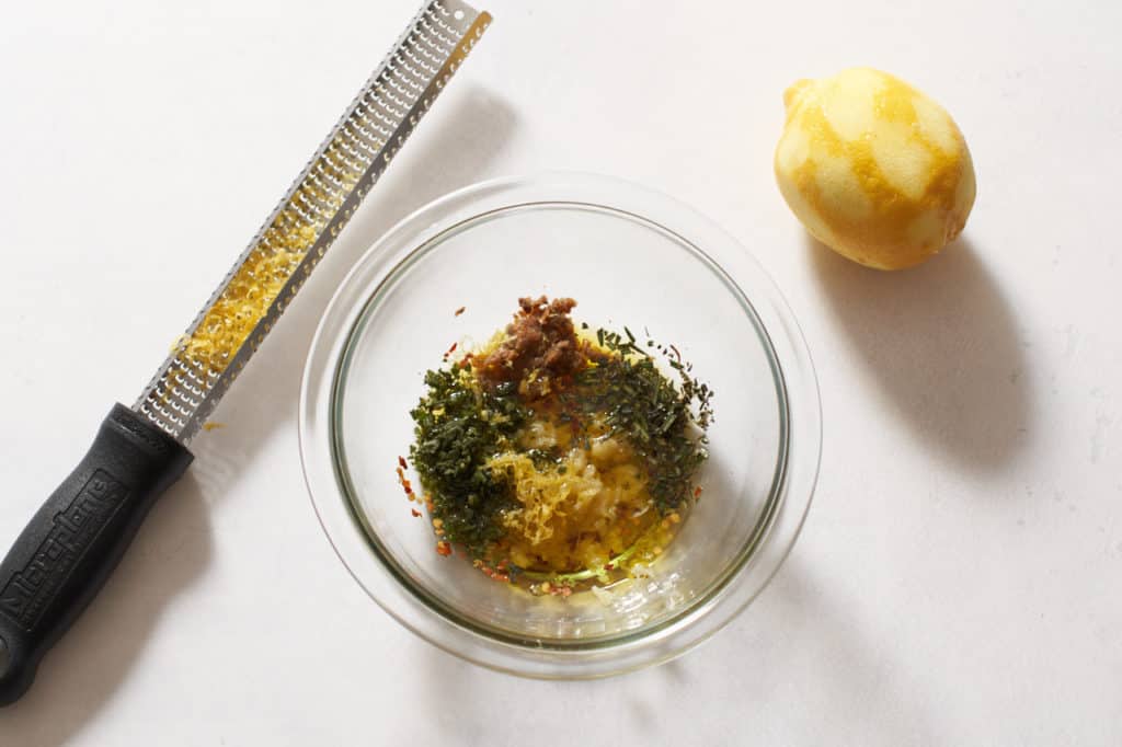 A microplane grater, a zested lemon, and a bowl with herbs, olive oil and anchovies.