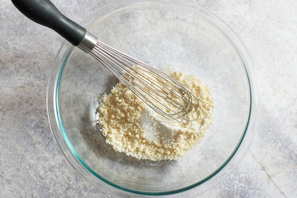 A whisk in a glass bowl of almond flour, salt, and baking powder.