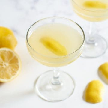 Two bee's knees cocktails in coupe glasses with two twists of lemon peel and cut fresh lemons..