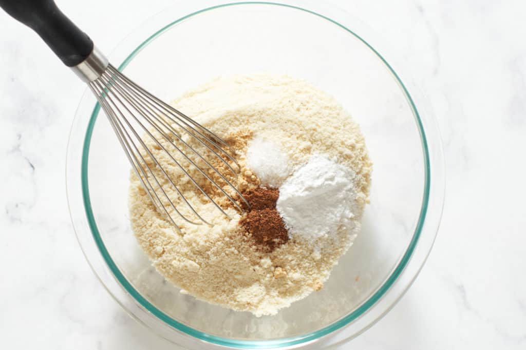 A whisk in a bowl full of almond flour with salt, ginger, nutmeg and baking powder.
