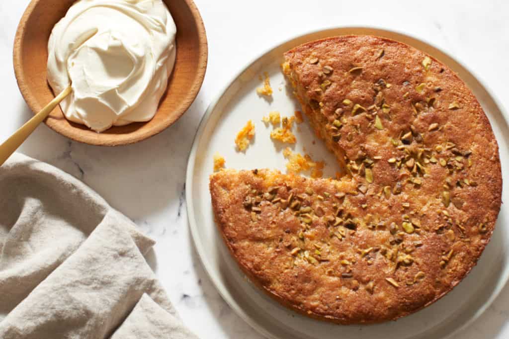 A gluten free carrot cake with a slice cut out of it next to a bowl of mascarpone cheese.