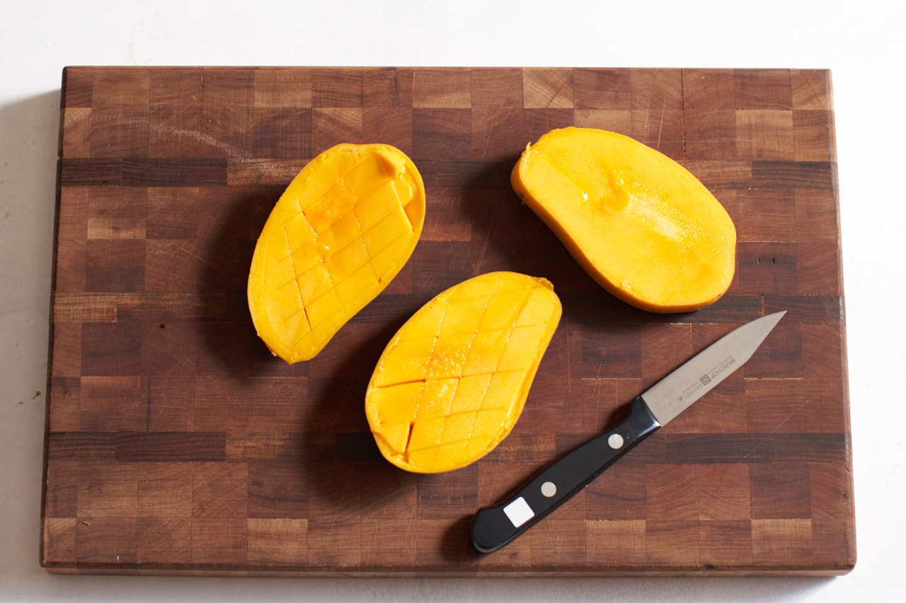 A mango slice in half with its seed on a cutting board with a paring knife.