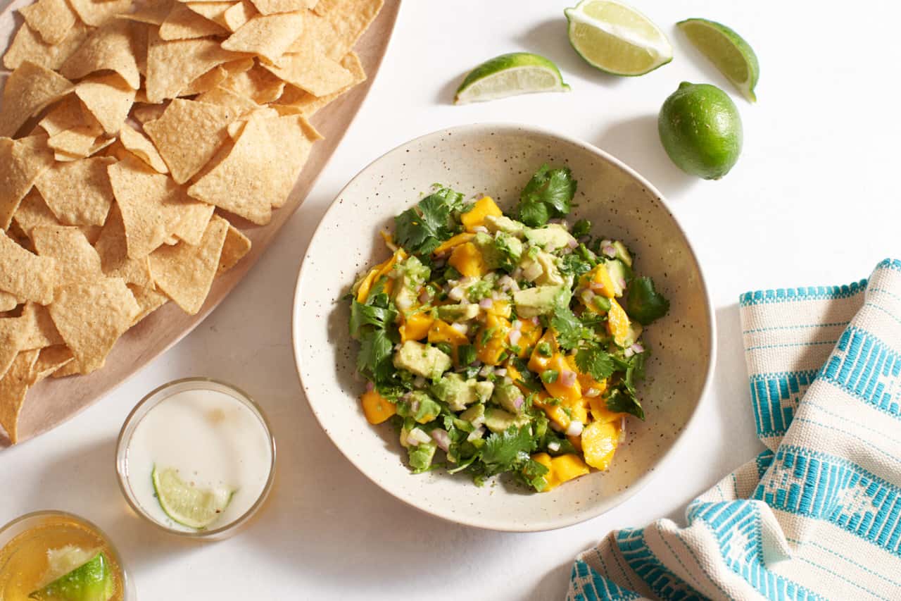 A bowl of mango salsa surrounded by a tray of corn chips, two glasses of beer, cut limes, and a turquoise and white towel with a Mexican pattern on it.