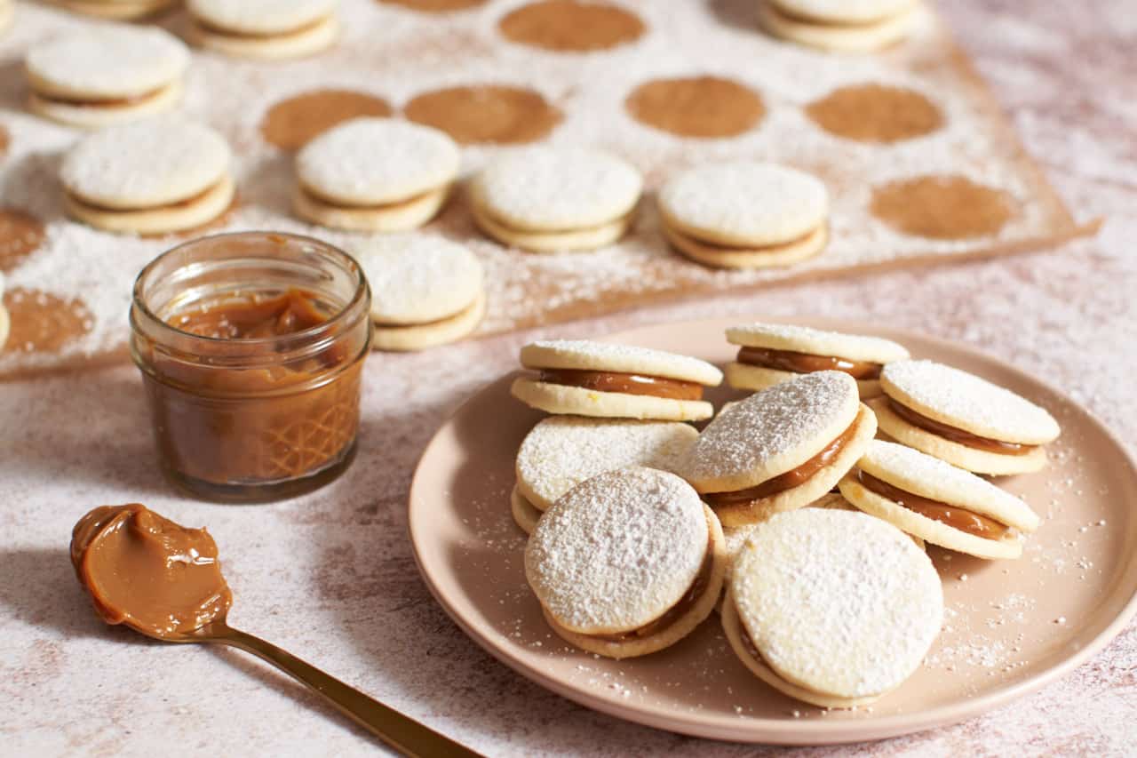 Alfajores stacked on a pink plate. A small jar and spoonful of dulce de leche are on the left, cookies on parchment dusted in powdered sugar are in the background.
