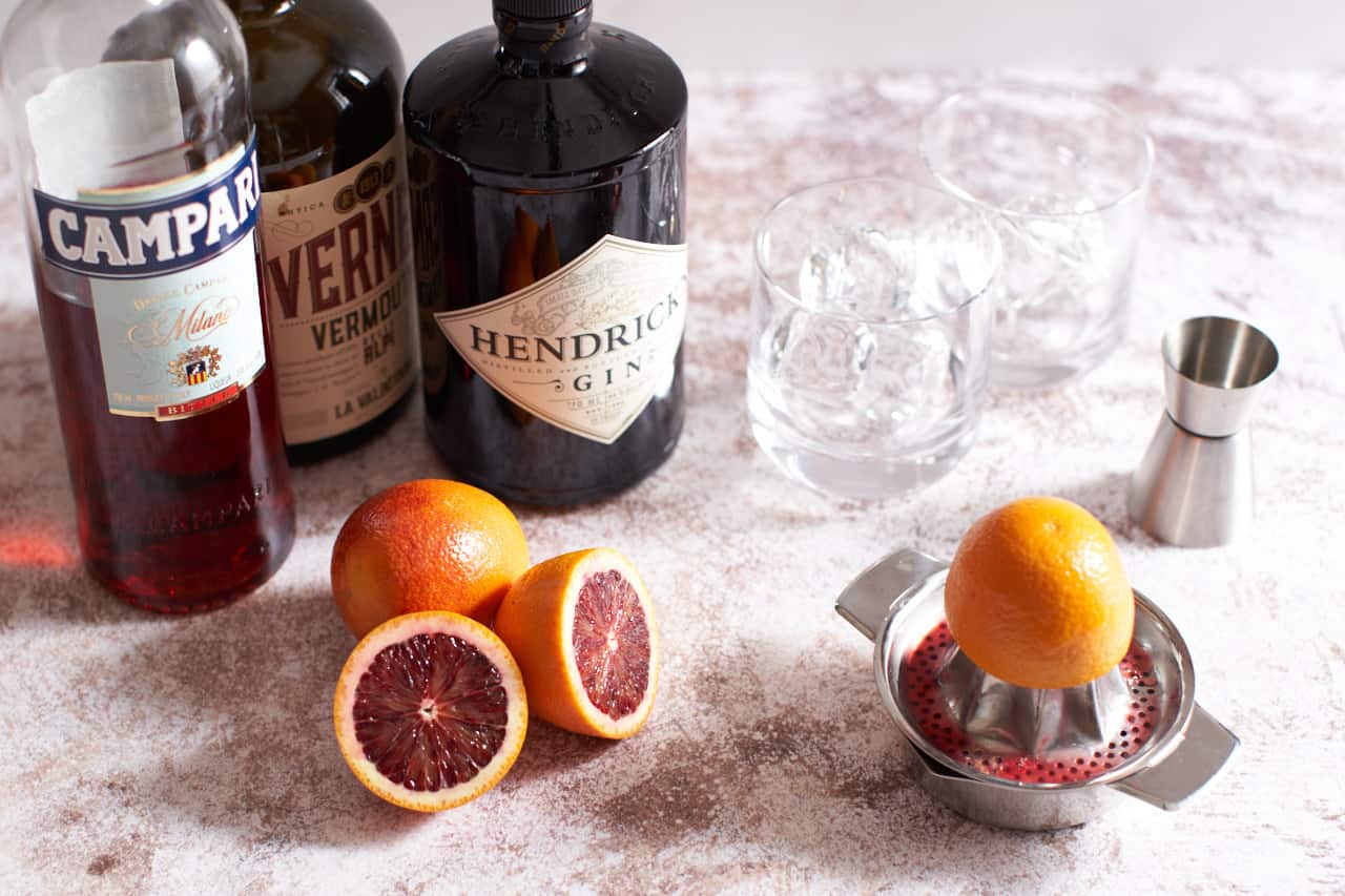 Bottles of Campari, vermouth, and gin, with sliced blood oranges, a juicer, a jigger, and cocktail glasses filled with ice.