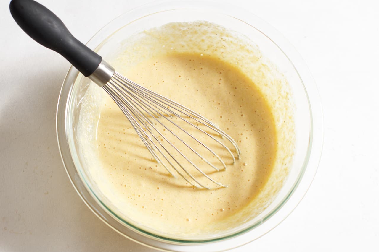 A whisk in a bowl of finished cornmeal batter that has begun to bubble as the rising agents activate.