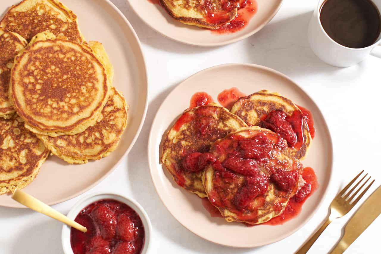 A plate of cornmeal pancakes topped with strawberry compote surrounded by a bowl of compote, a large serving plate of pancakes, a cup of coffee, and gold-colored utensils.