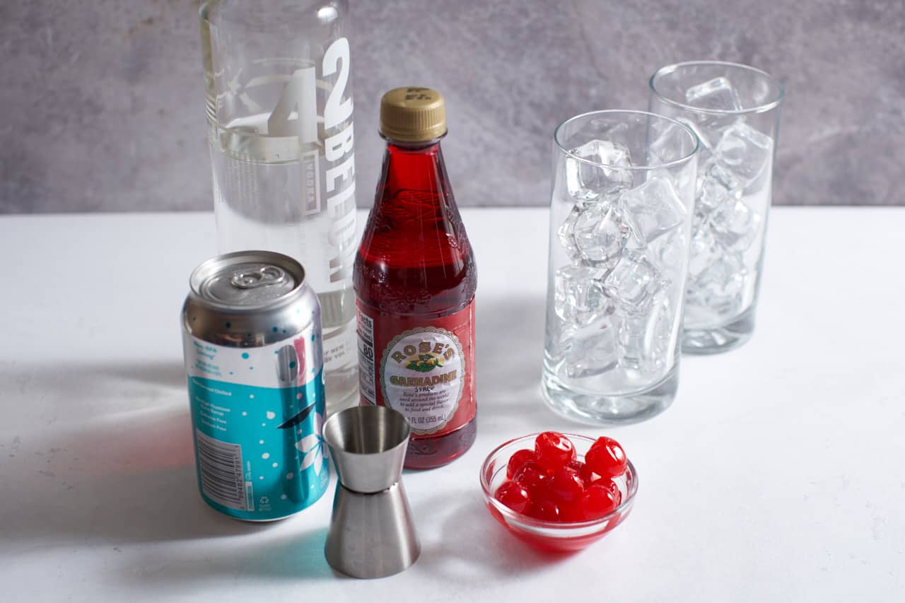 A can of soda, bottles of vodka and grenadine, a cocktail jigger, a bowl of maraschino cherries and two high ball glasses full of ice.