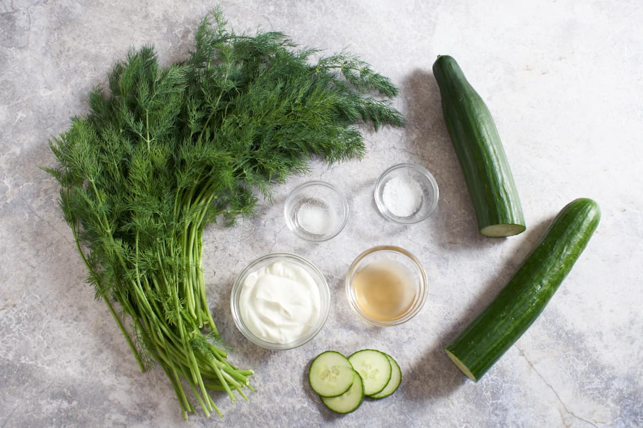 Fresh dill, an English cucumber sliced in half, and small glass bowls of sour cream, salt, sugar, and cider vinegar.