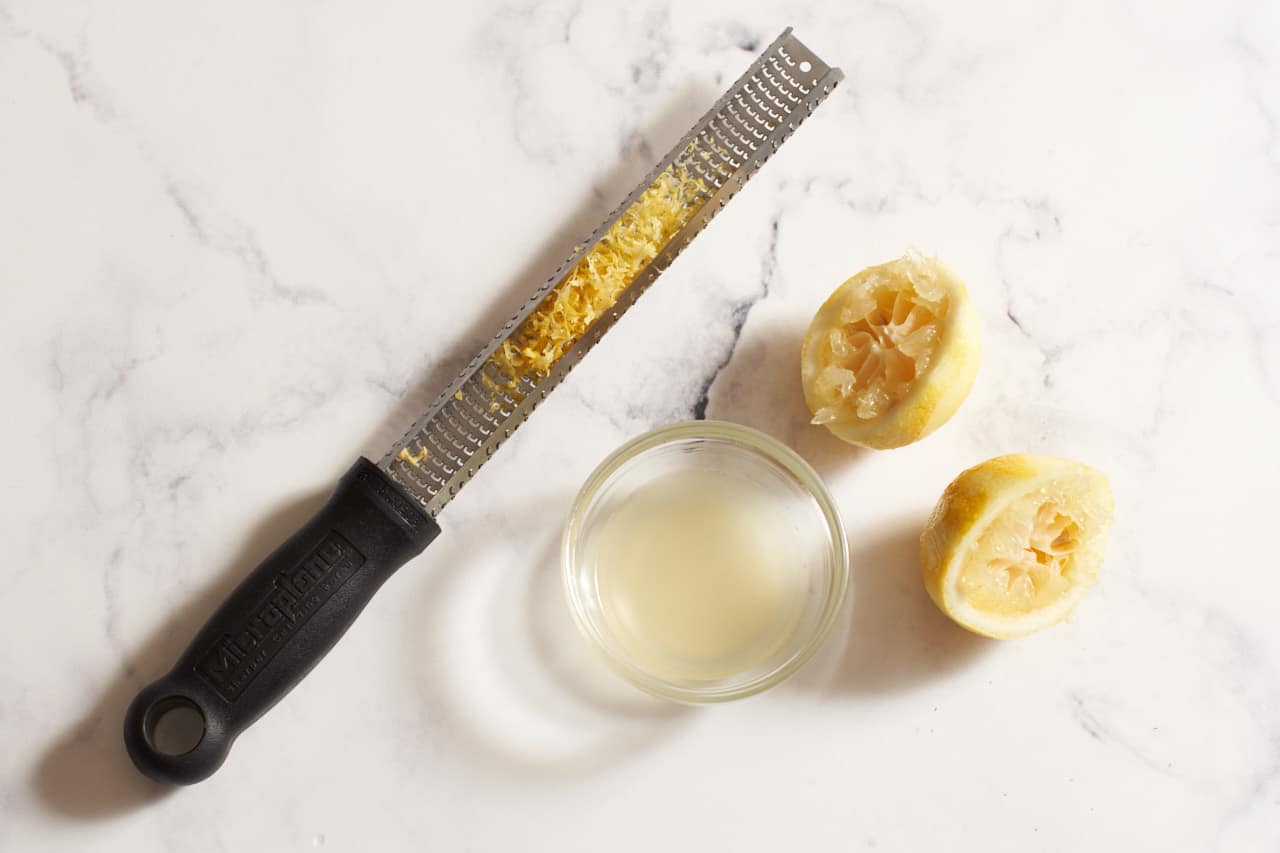 A microplane grater with zest next to a bowl of lemon juice and two squeezed lemon halves.
