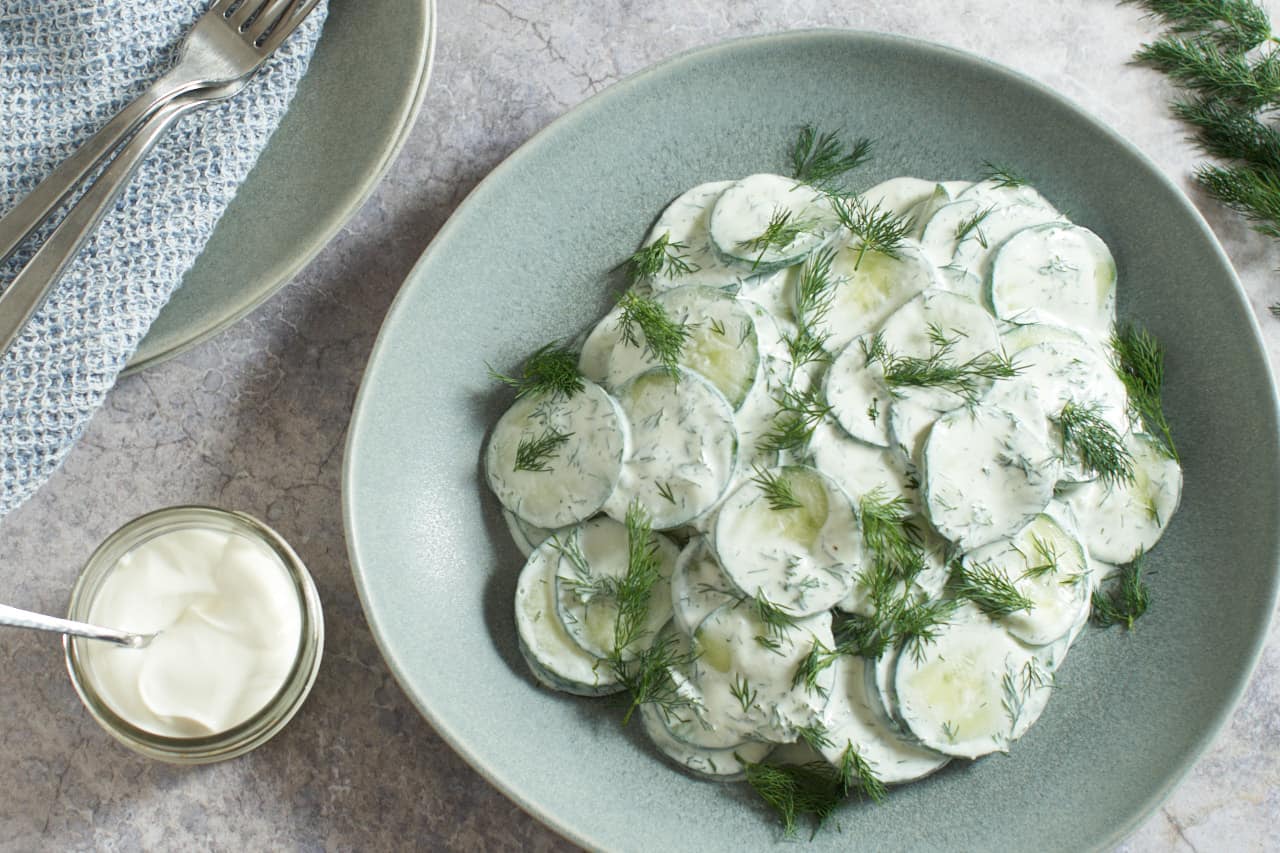 Creamy cucumber salad in a gray bowl. Two plates stacked with a blue napkin and two forks, and a small glass jar of sour cream are on the left, fresh dill is in the top right corner.