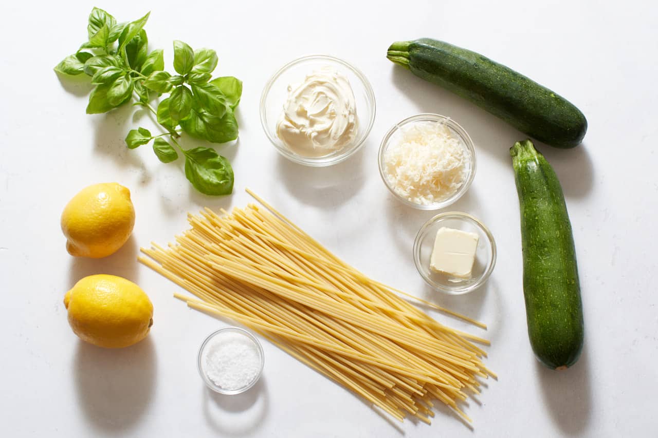 Lemons, zucchini, fresh basil, and linguine pasta noodles next to small glass bowls of mascarpone cream cheese, parmesan, butter, and salt.