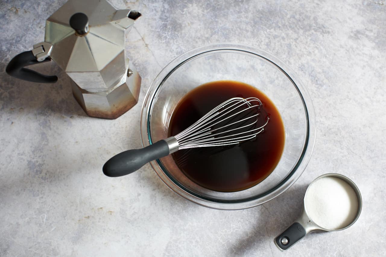 A moka pot, a glass bowl of coffee with a whisk in it, and a measuring cup of sugar.