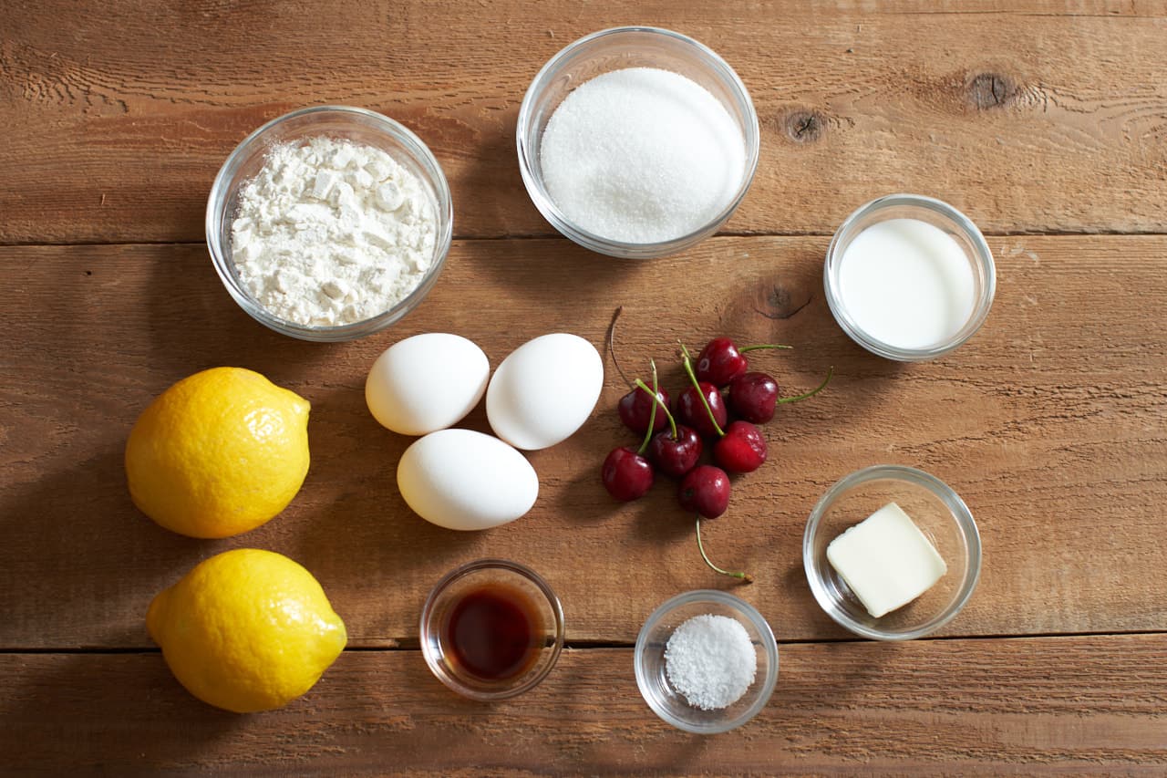 Lemons, charries, and eggs surrounded by small bowls of flour, sugar, milk, salt, butter and vanilla.