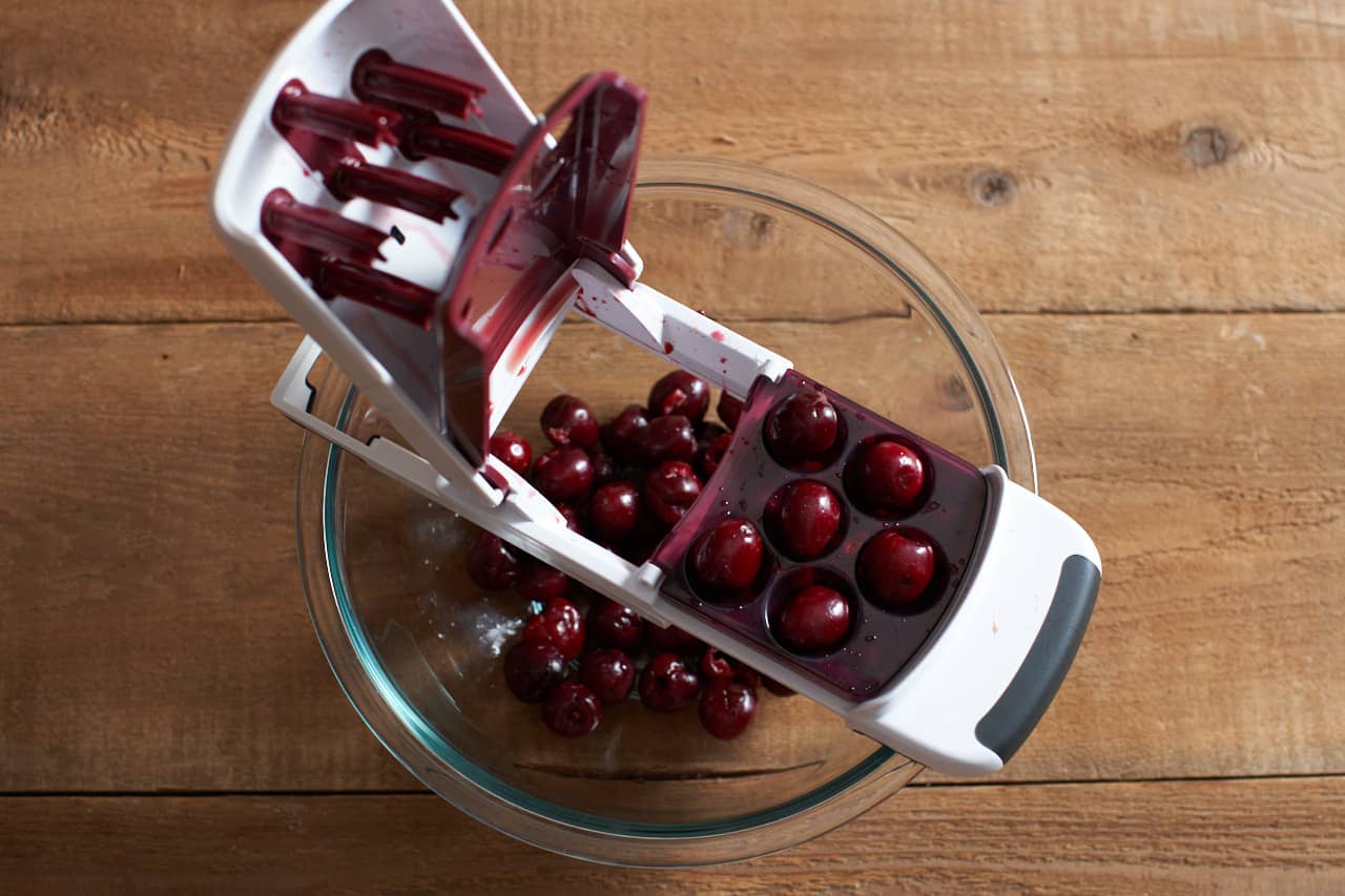 A cherry pitting tool over a glass bowl full of pitted fresh cherries.