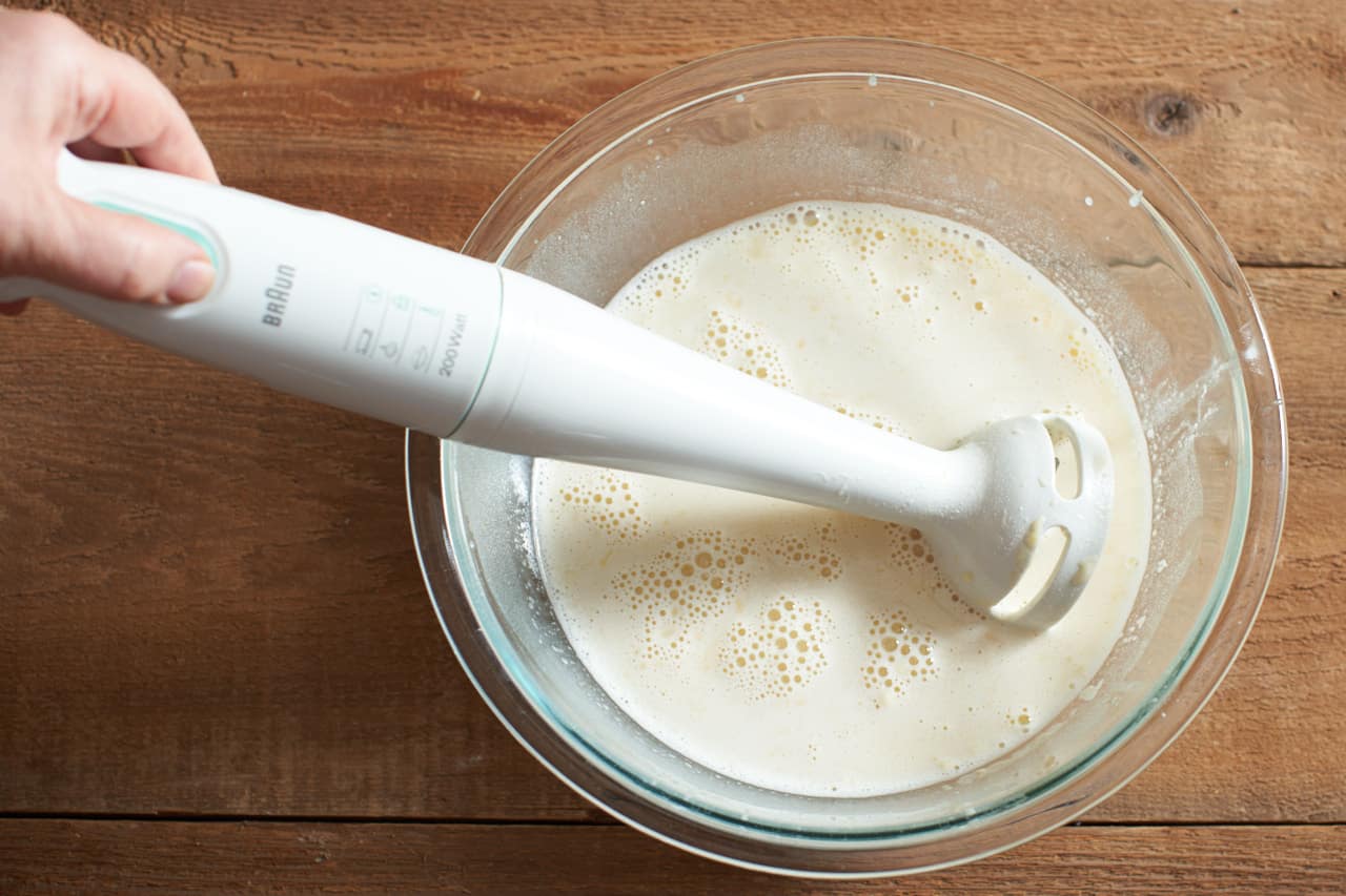 A woman's hand holding an emulsion blender in a bowl of clafoutis batter.