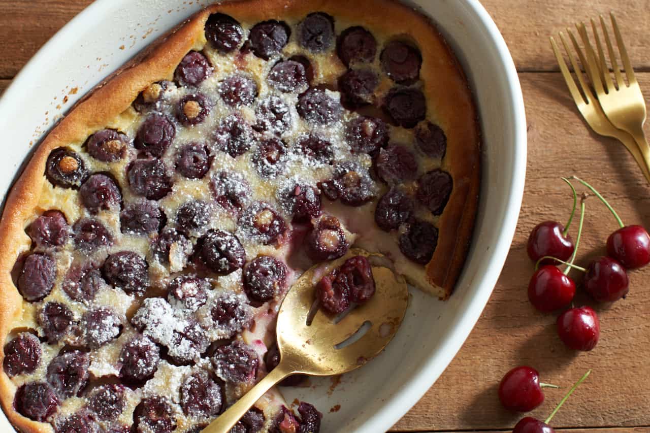 Cherry clafoutis in a white oval baking dish. A gold spoon rests in a spot where a serving has been scooped out. Fresh cherries and two gold forks are on the right.