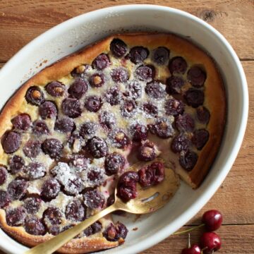 Cherry clafoutis in an oval baking dish with a gold spoon. A few fresh cherries are on the table in the bottom right corner.