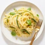 Creamy zucchini pasta topped with fresh basil and lemon zest on a white plate with a gold fork.